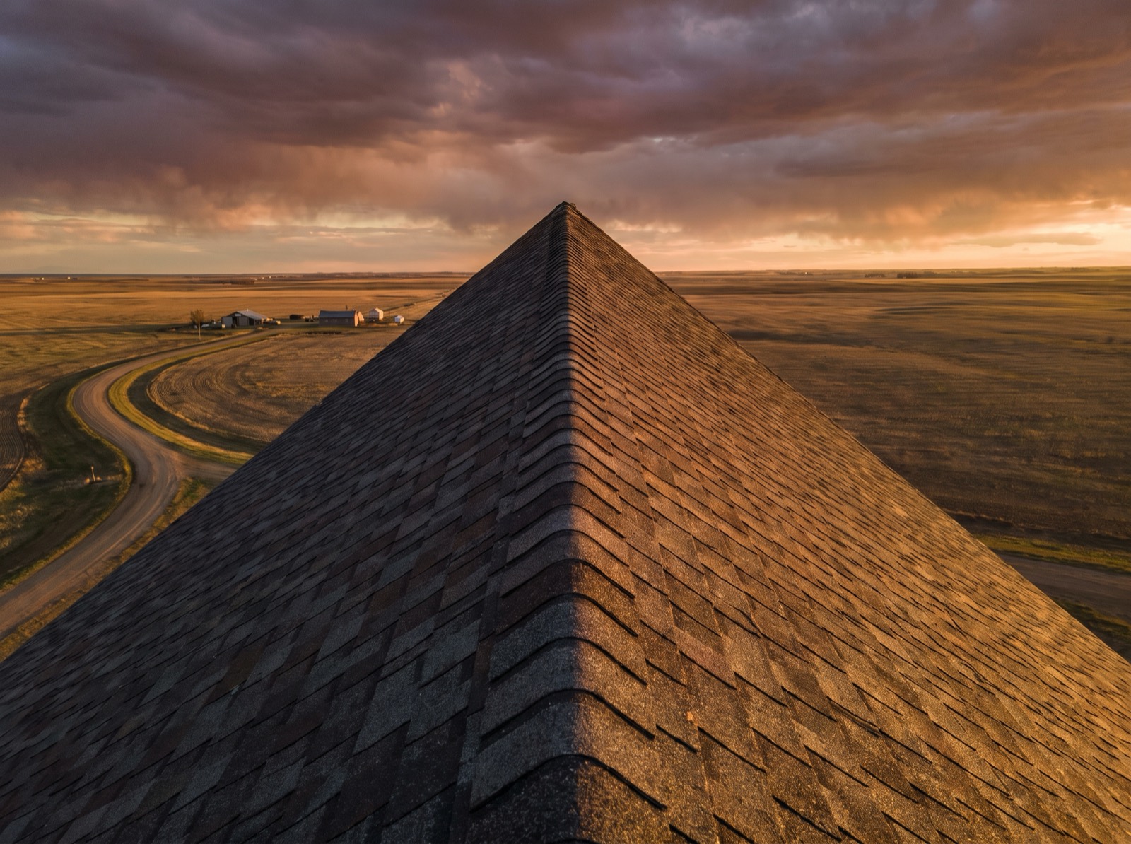 A residential shingle roof seen from the ridge at sunset — one plane in warm light, one in shadow — over the Alberta prairie.